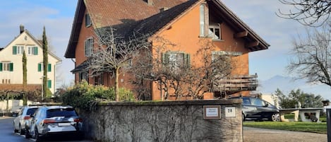A two-story building with a sloped roof, a stone wall in front, and parked cars.
