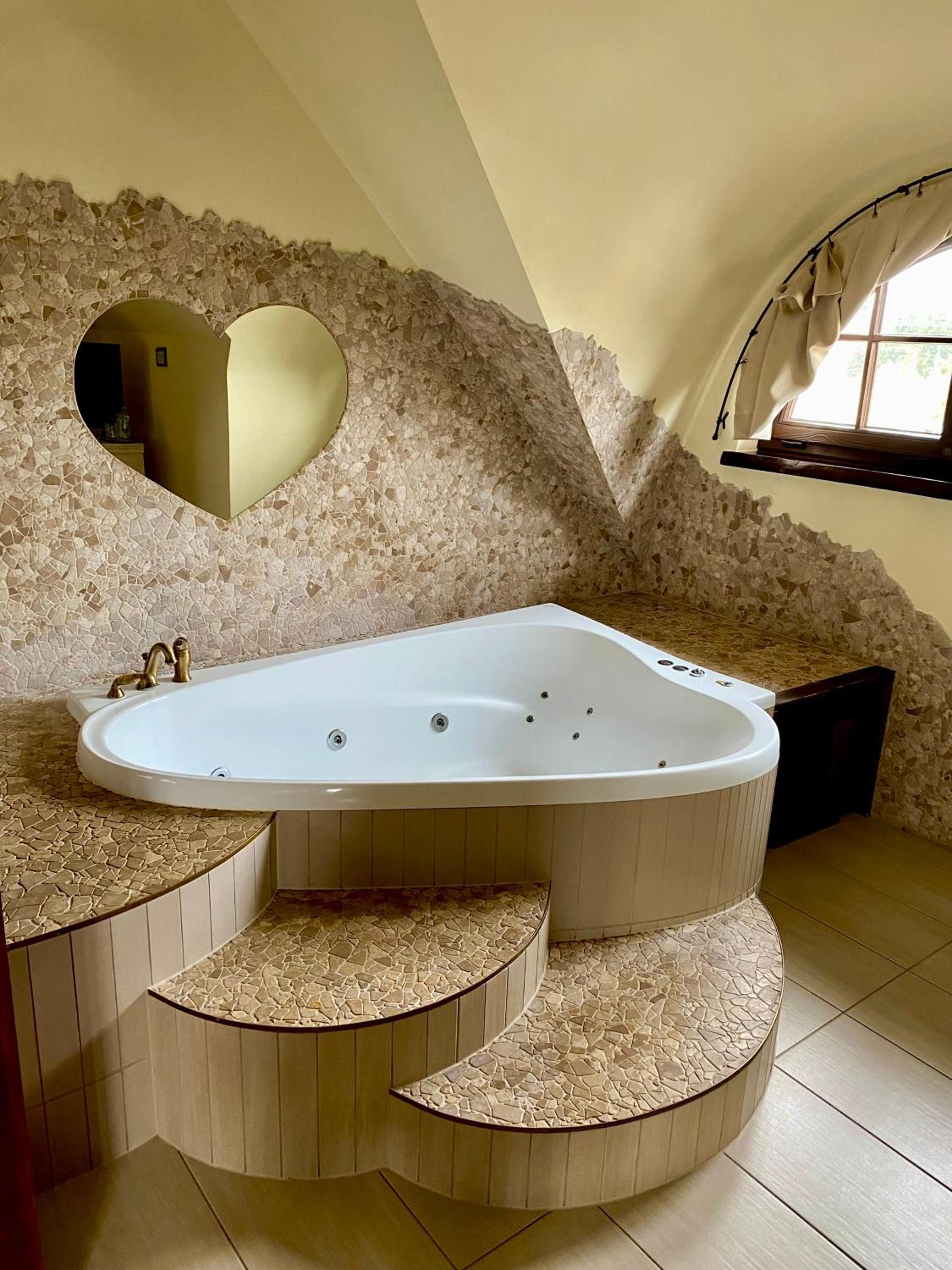Un baño moderno con una bañera independiente, paredes de mosaico de piedra y un ventanal con cortinas.