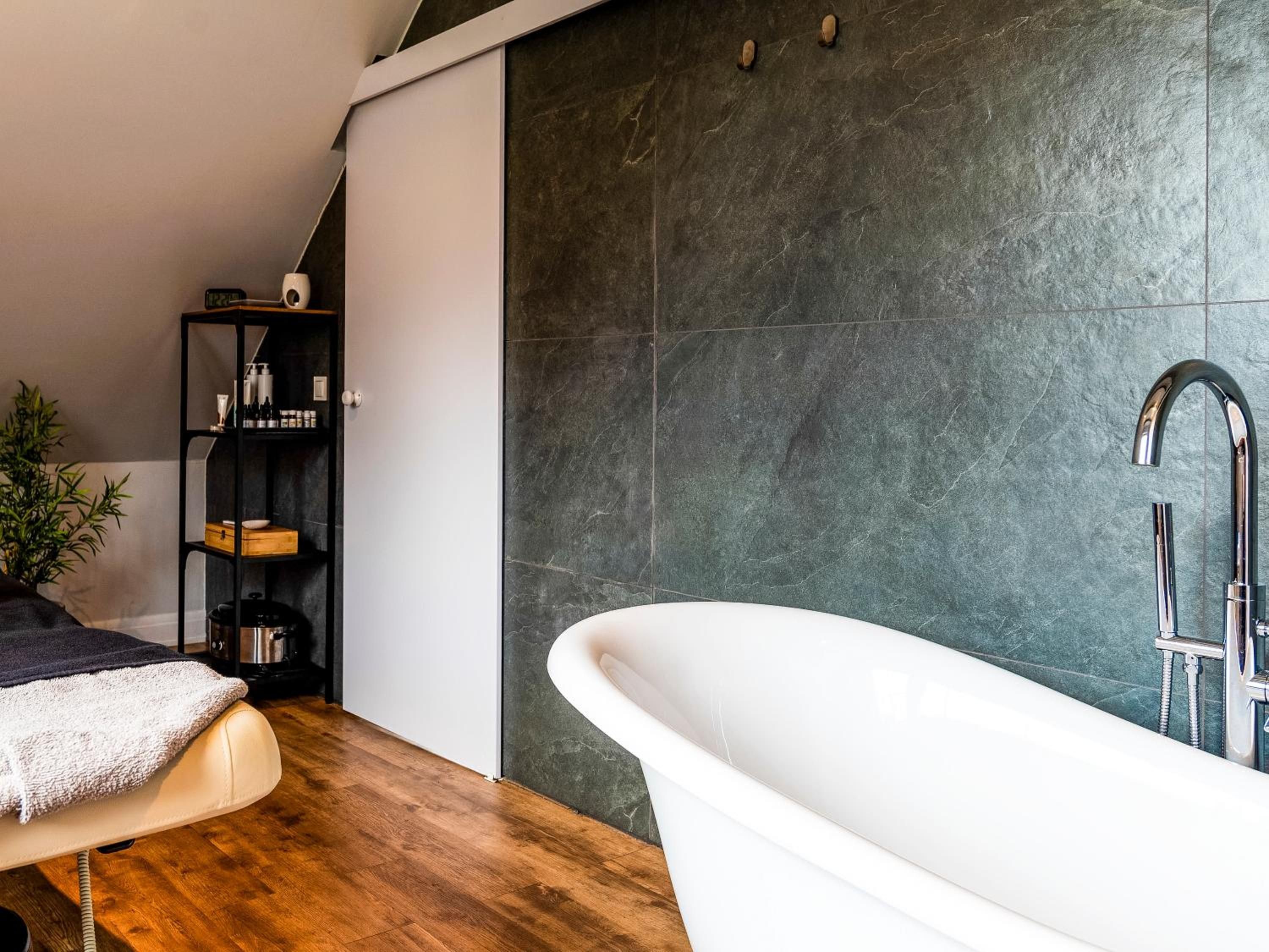 A modern bathroom with a freestanding bathtub, dark stone wall, and a wooden floor.
