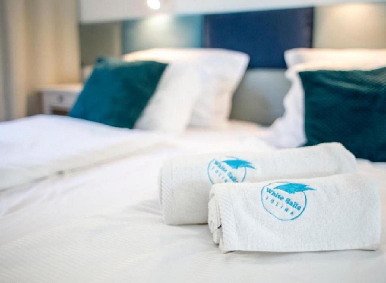 Ein Hotelzimmer mit zwei Betten, jeweils mit weißer Bettwäsche und türkisfarbenen Kissen. Es liegen zwei gefaltete Handtücher mit einem Logo darauf.