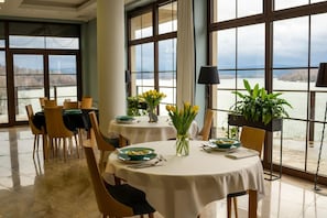 A dining area with tables set for two, large windows with a view of a body of water, and a potted plant on the windowsill.