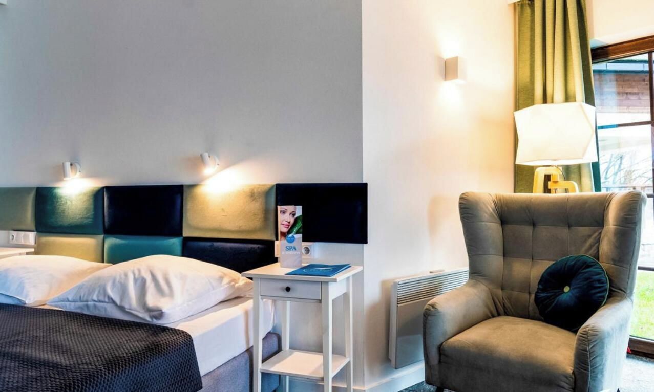 A hotel room with a bed, a bedside table, a chair, a lamp, and a window with curtains.