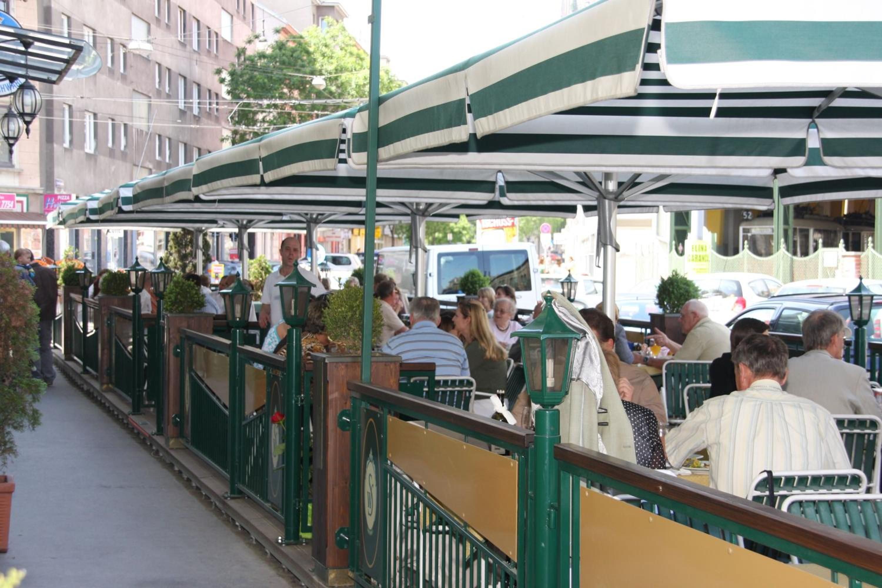 Un caffè all'aperto con tavoli e sedie sotto una tenda a righe verdi e bianche, circondato da edifici e da una strada.