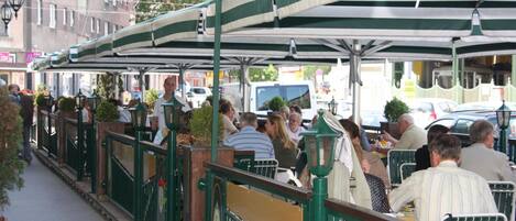 An outdoor café with tables and chairs under a green and white striped awning, surrounded by buildings and a street.