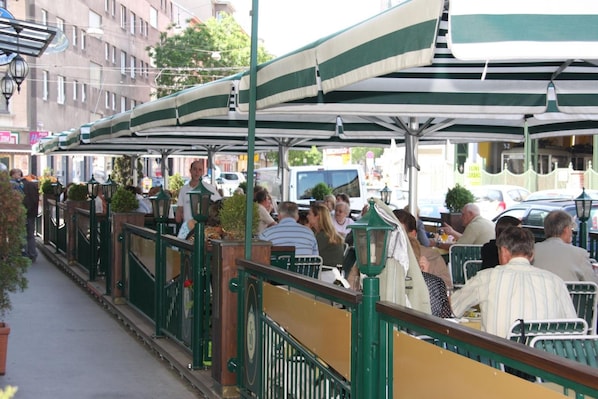 Un café al aire libre con mesas y sillas bajo un toldo verde y blanco a rayas, rodeado de edificios y una calle.