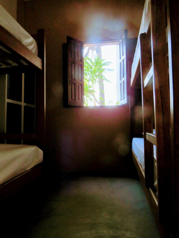 A narrow room with bunk beds, a window with open shutters, and a view of greenery outside.