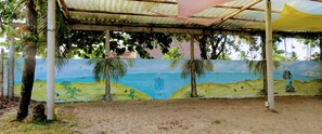 A covered outdoor area with a mural depicting a beach scene, palm trees, and a lighthouse. - Tribo De Gaia (Conceicao da Barra)