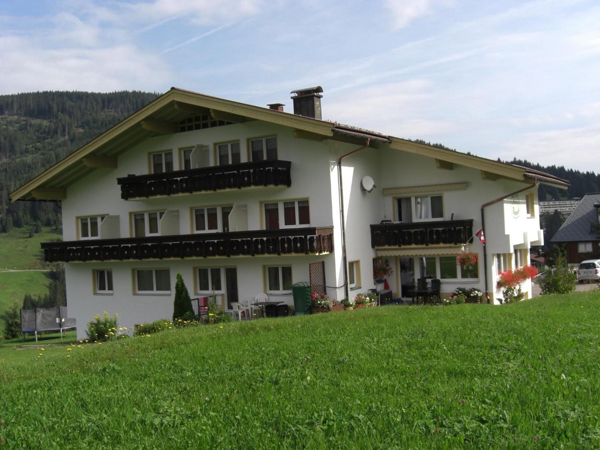 A two-story building with a balcony on the upper floor, surrounded by a green field and a forested hill in the background.