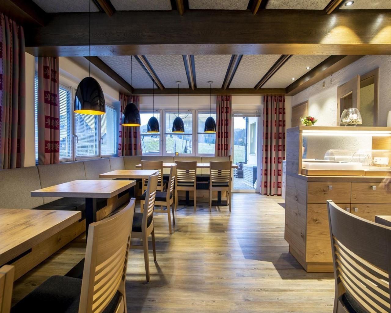 Interior de restaurante con mesas y sillas de madera, iluminación colgante y una barra con pileta.
