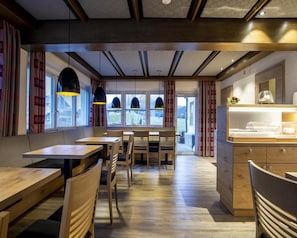 Interior de un restaurante con mesas y sillas de madera, iluminación colgante y vistas amplias a través de grandes ventanales.