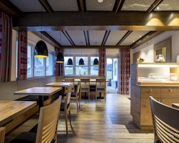 A restaurant interior with wooden tables and chairs, pendant lighting, and a view through large windows.