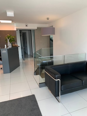 A modern lobby with a reception desk, a black leather sofa, a glass railing, and a staircase. - Haus 7 - Boardinghouse (Kornwestheim)