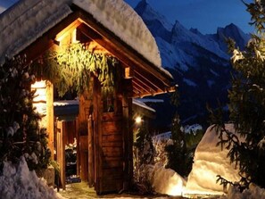 Una cabaña de madera con techo nevado, rodeada de nieve y montañas.