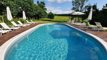 A rectangular swimming pool with clear blue water, surrounded by white lounge chairs and umbrellas, set against a backdrop of trees and a clear sky.