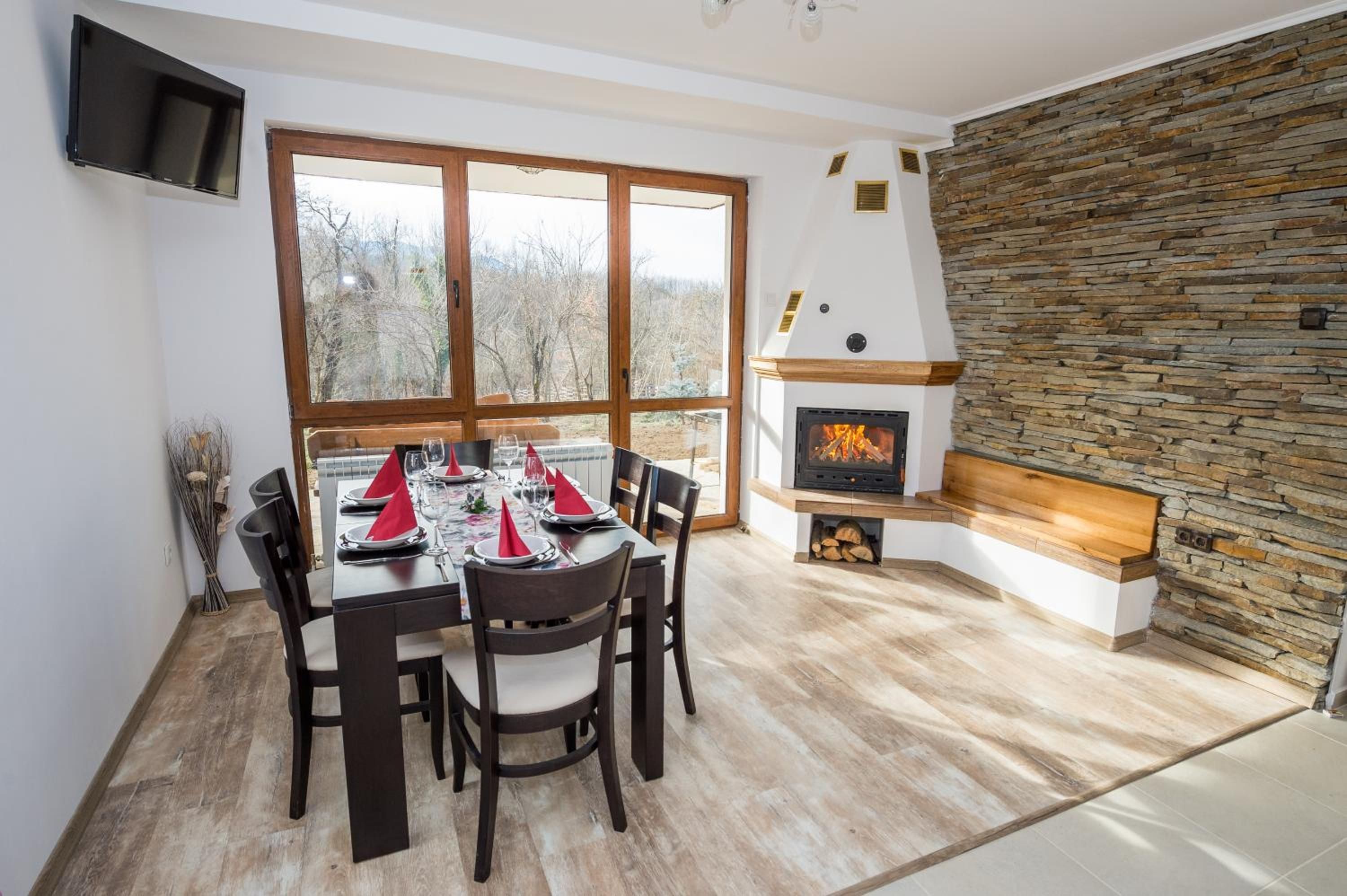 A dining area with a table set for four, a stone fireplace, and a large window with a view of trees.