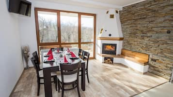 A dining area with a table set for four, a stone fireplace, and a large window with a view of trees.