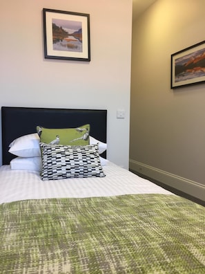 Cama bem arrumada com colcha verde estampada, dois travesseiros e uma pintura emoldurada de paisagem acima.