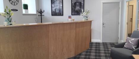 A reception area with a wooden counter, a black and white checkered floor, a grey sofa, and a door in the background.
