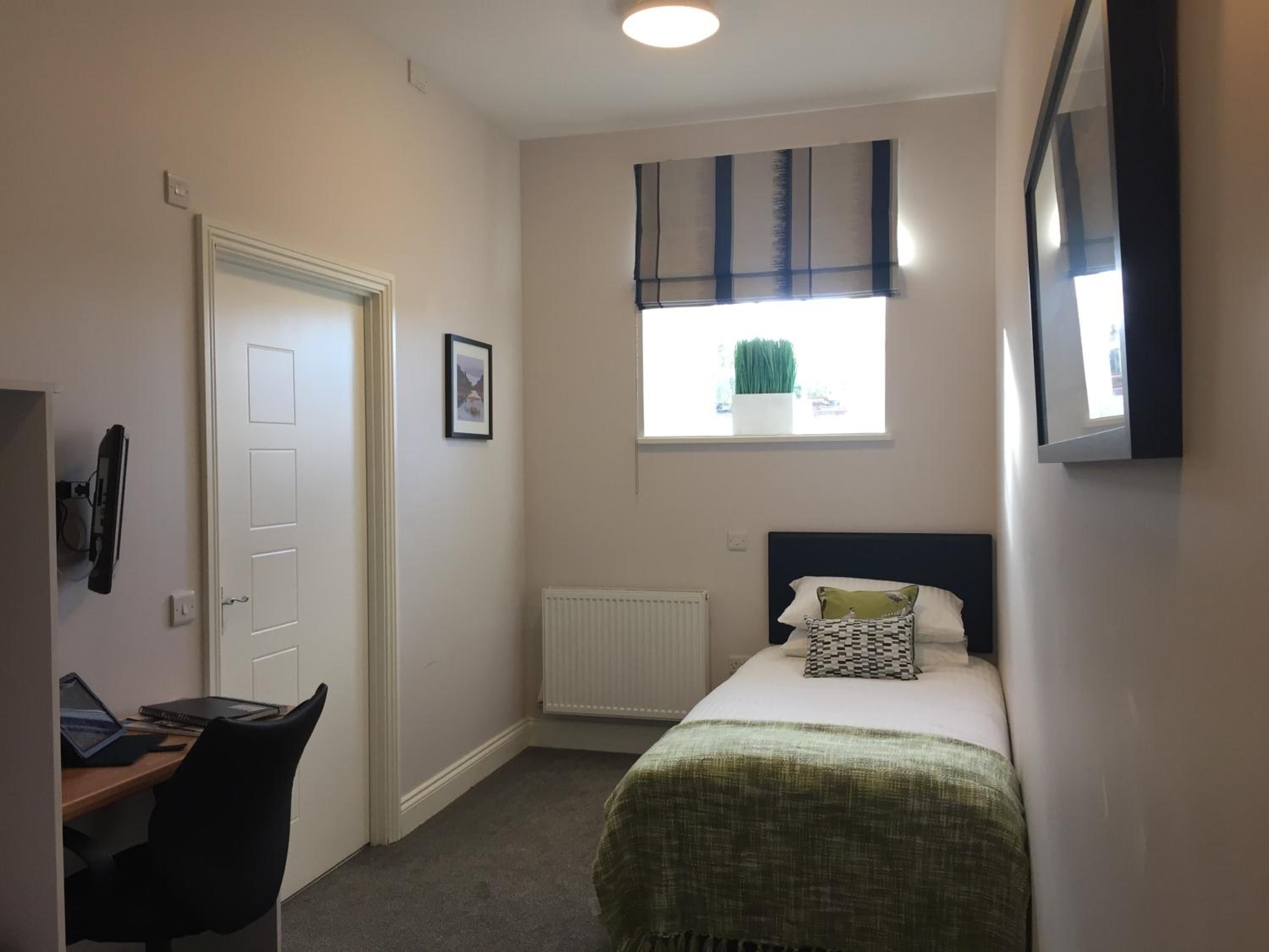 A compact bedroom with a single bed, a desk with a laptop, a radiator, and a window with a plant.