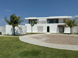 Uma casa moderna de dois andares com um acesso pavimentado, paredes brancas e palmeiras.
