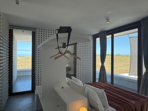 Quarto moderno com cama, luminária pendente e vista para a praia.