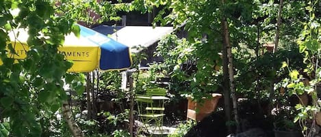 Un espacio al aire libre con sillas y mesas de metal, rodeado de follaje y un muro de piedra.