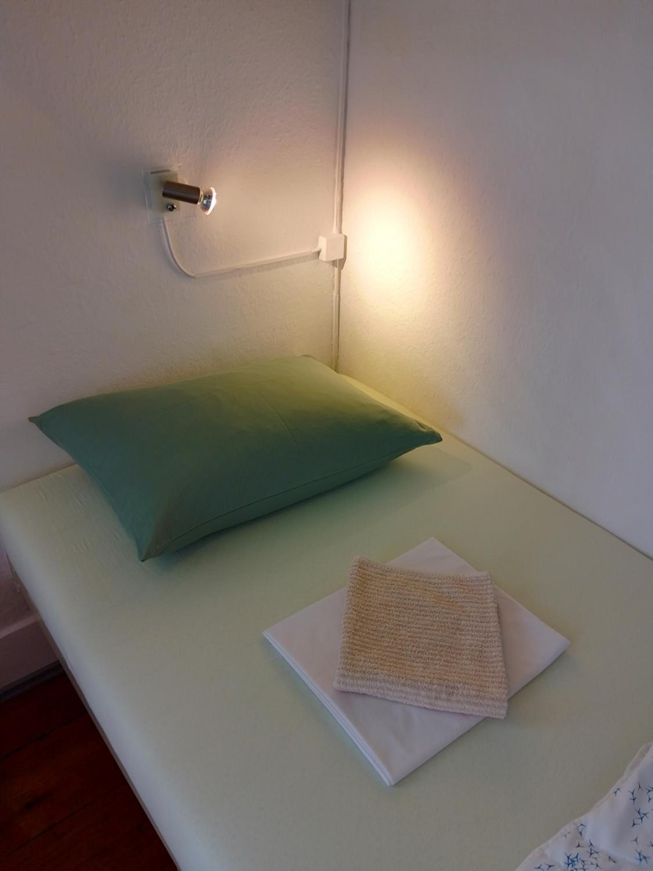 A small, single-bed room with a green pillow and a folded towel on the bed.