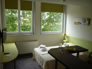 A room with two beds, a table with a lamp, a window with a view of trees, and a radiator. - West Appart' Hôtel (Bessines)