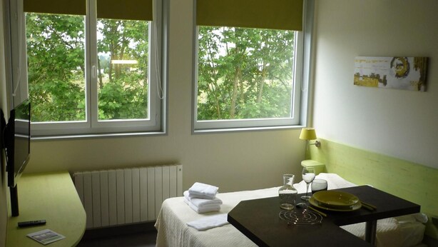 A room with two beds, a table with a lamp, a window with a view of trees, and a radiator. - West Appart' Hôtel (Bessines)