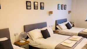 A hotel room with three beds, each with a pillow, a bedside table with a lamp, and a wall-mounted fan. - The Players Golf Club (Bristol)
