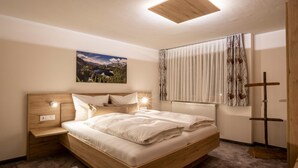 Un dormitorio con cama de madera, mesita de noche, lámpara de pared, ventana con cortinas translúcidas y un cuadro de paisaje.