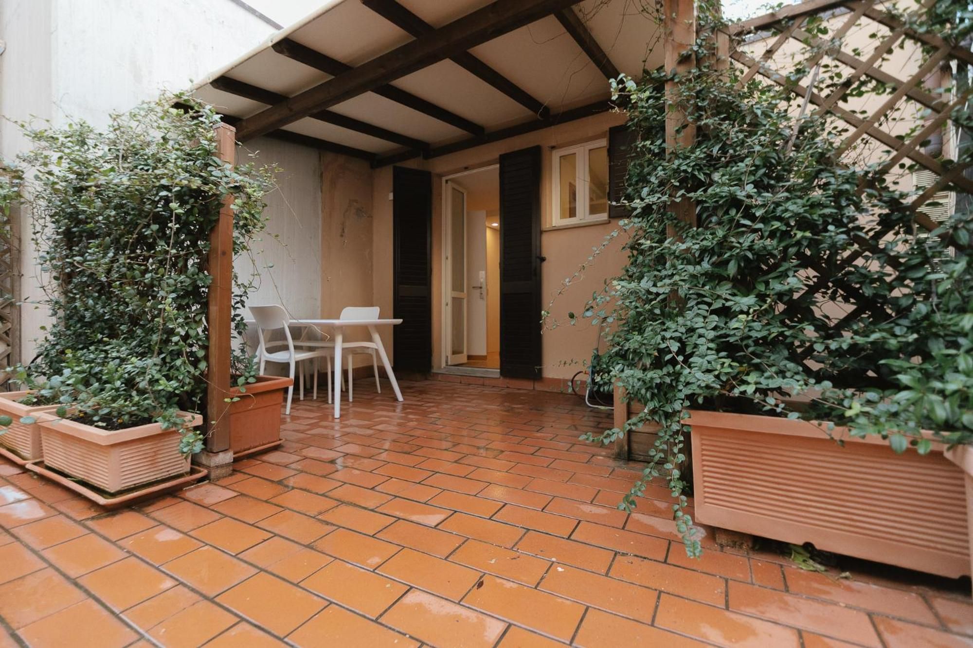 Eine überdachte Terrasse mit Terrakottafliesen, einem weißen Tisch und Stühlen sowie Blumentöpfen.