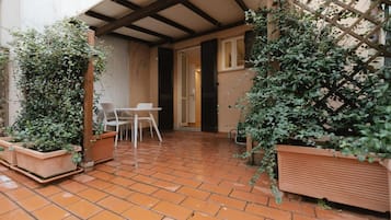 Eine überdachte Terrasse mit Terrakottafliesen, einem weißen Tisch und Stühlen sowie Blumentöpfen.
