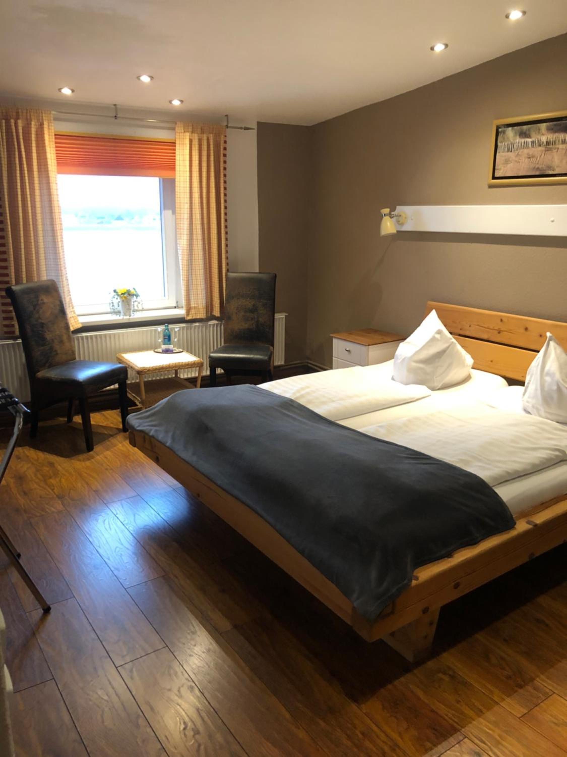 Ein Hotelzimmer mit einem hölzernen Bett, einem Sessel, einem kleinen Tisch, einem Heizkörper und einem Fenster mit Vorhängen.