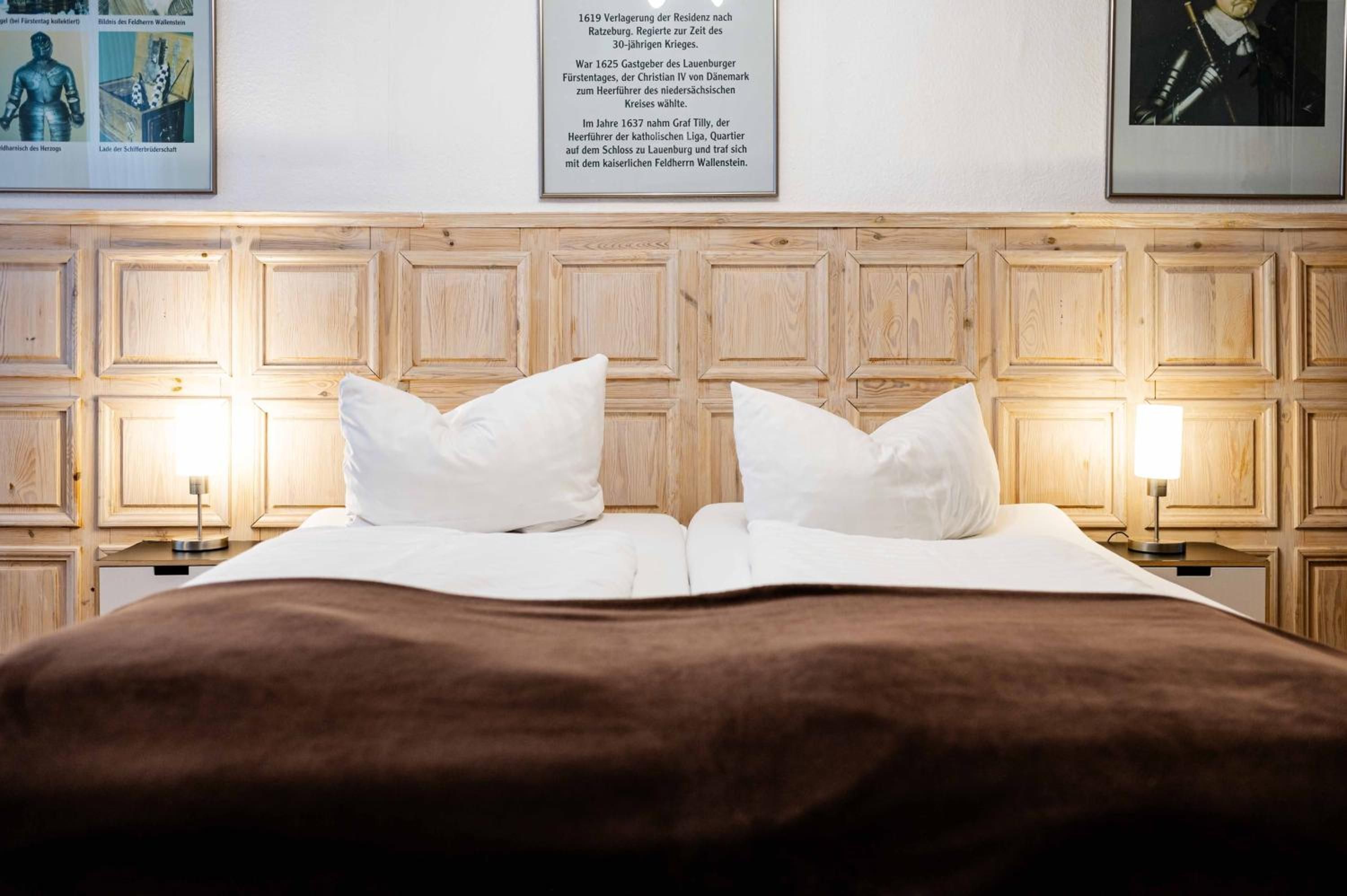 Een hotelkamer met een houten hoofdeinde, twee witte kussens, een bruine deken en twee nachtlampjes.