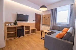 Una sala moderna con un sofá gris, estanterías de madera, un televisor de pantalla plana, un pequeño árbol de Navidad y una ventana con cortinas.