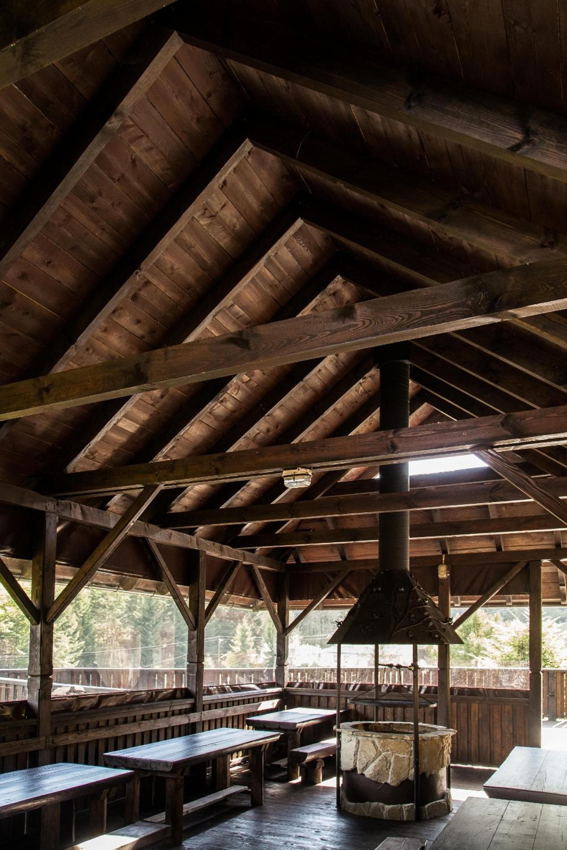 Una estructura rústica de madera con techo alto y chimenea central. Hay bancos y mesas de madera dispuestas para sentarse.