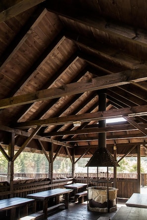 Una estructura rústica de madera con techo alto y chimenea central. Hay bancos y mesas de madera dispuestas para sentarse.
