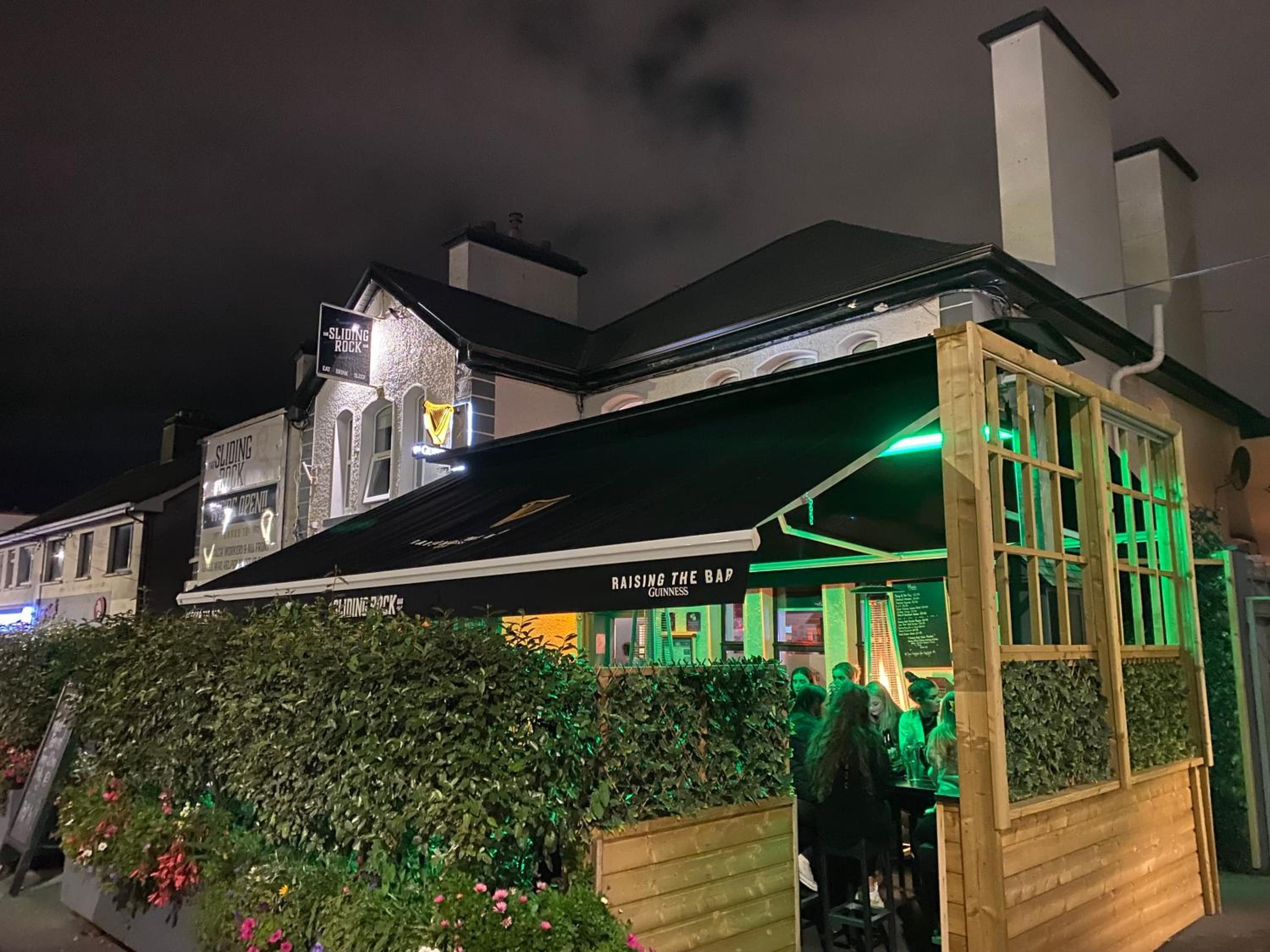 A bar with a green-lit exterior and a sign that reads 'RAISING THE BAR GUINNESS.'