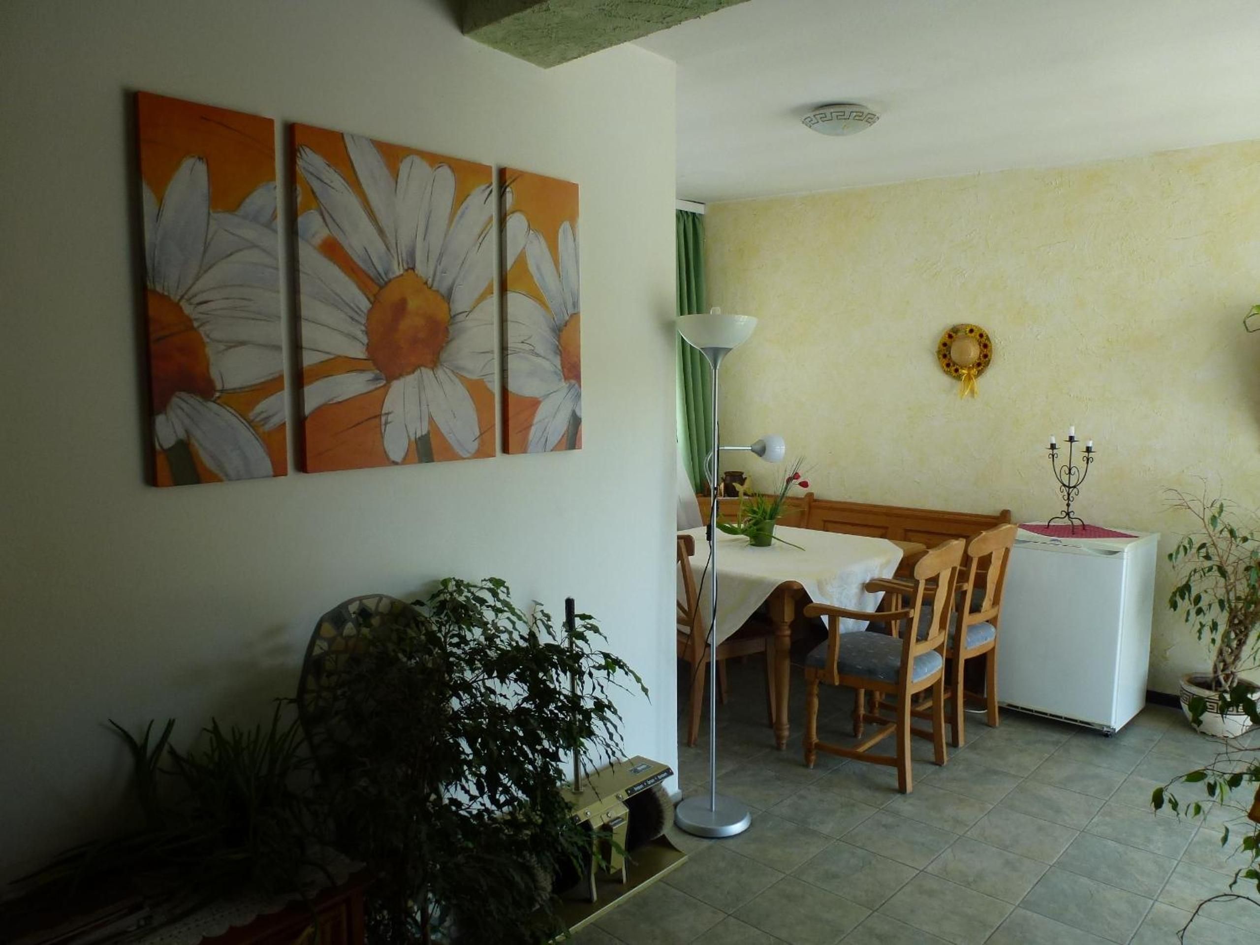 Ein Essbereich mit Tisch, Stühlen, einer Stehlampe und einem Kühlschrank. An der Wand hängen zwei Bilder und im Vordergrund steht eine Topfpflanze.