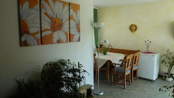 Ein Essbereich mit Tisch, Stühlen, einer Stehlampe und einem Kühlschrank. An der Wand hängen zwei Bilder und im Vordergrund steht eine Topfpflanze.