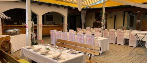 Un área de comedor al aire libre con mesas dispuestas para una comida, cubierta por un toldo a rayas y rodeada de bancos de madera.