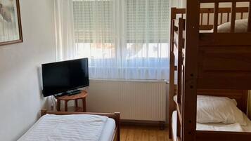 Ein Zimmer mit einem Einzelbett, einem Fernseher auf einem kleinen Tisch, einem Fenster mit durchsichtigen Vorhängen und einem hölzernen Etagenbett.