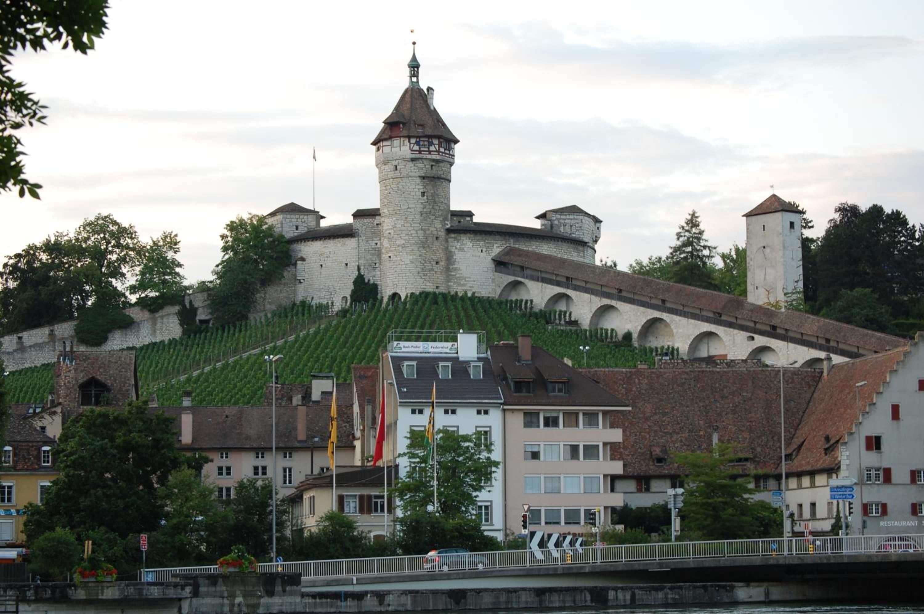 A historic castle with a prominent tower, surrounded by modern buildings and a river.