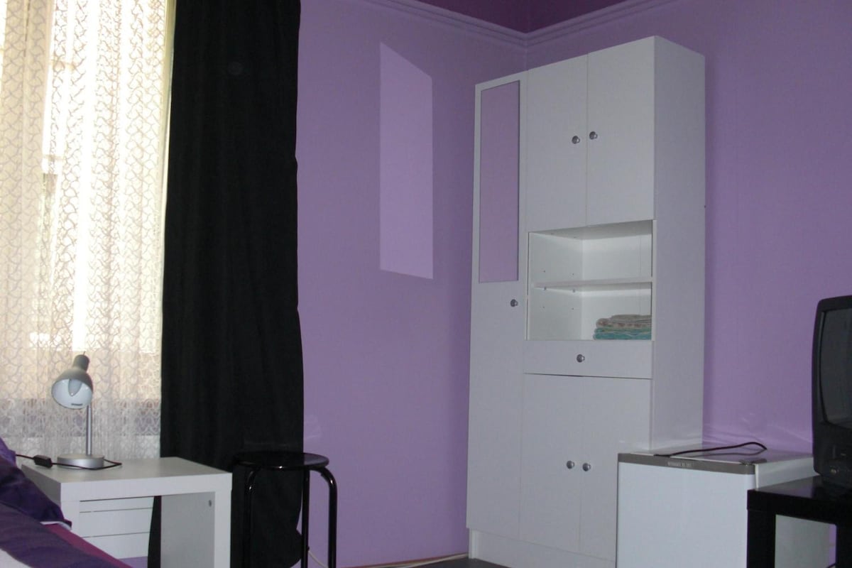 Ein Zimmer mit weißem Schreibtisch, schwarzem Stuhl, weißem Kleiderschrank und Fernseher auf einem Sockel.