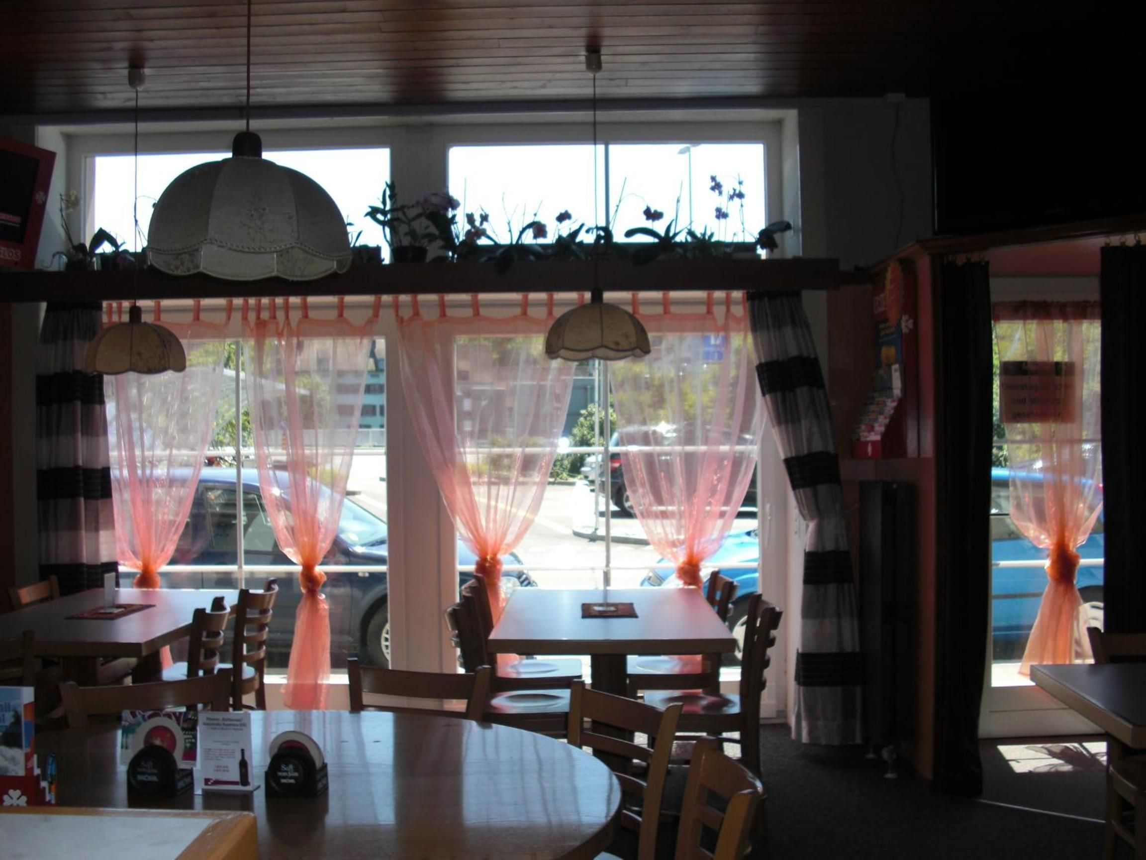 A restaurant interior with round tables, chairs, and a view of the street through large windows with sheer curtains.
