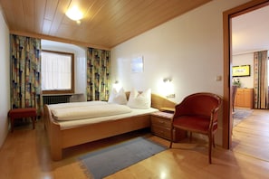 Ein Schlafzimmer mit Bett, Sessel, Nachttisch, Lampe, einem Fenster mit Vorhängen und einer Tür zu einem anderen Raum.
