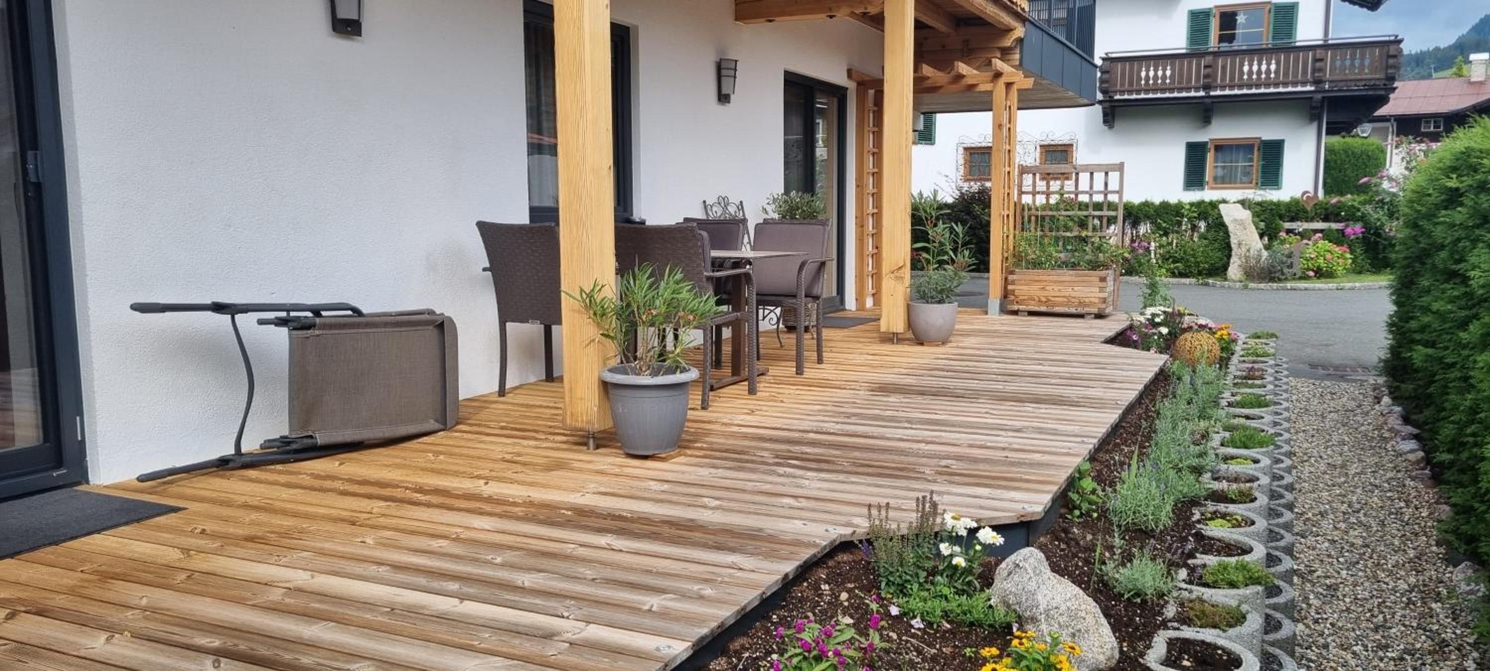 Um deck de madeira com móveis de exterior, plantas em vasos e um jardim com caminho de pedra.