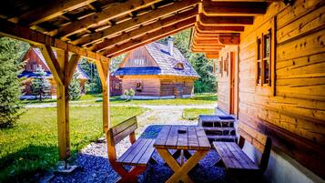 A wooden cabin with a covered porch, a picnic table, and a view of a traditional house in the background.