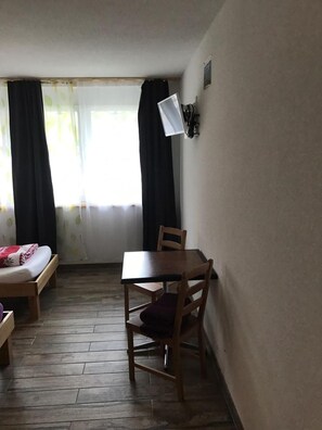 A room with a bed, a table, and a chair. There is a window with curtains, a wall-mounted lamp, and a wooden floor. - Swiss Chalet Holidays Ringgenberg (Ringgenberg)